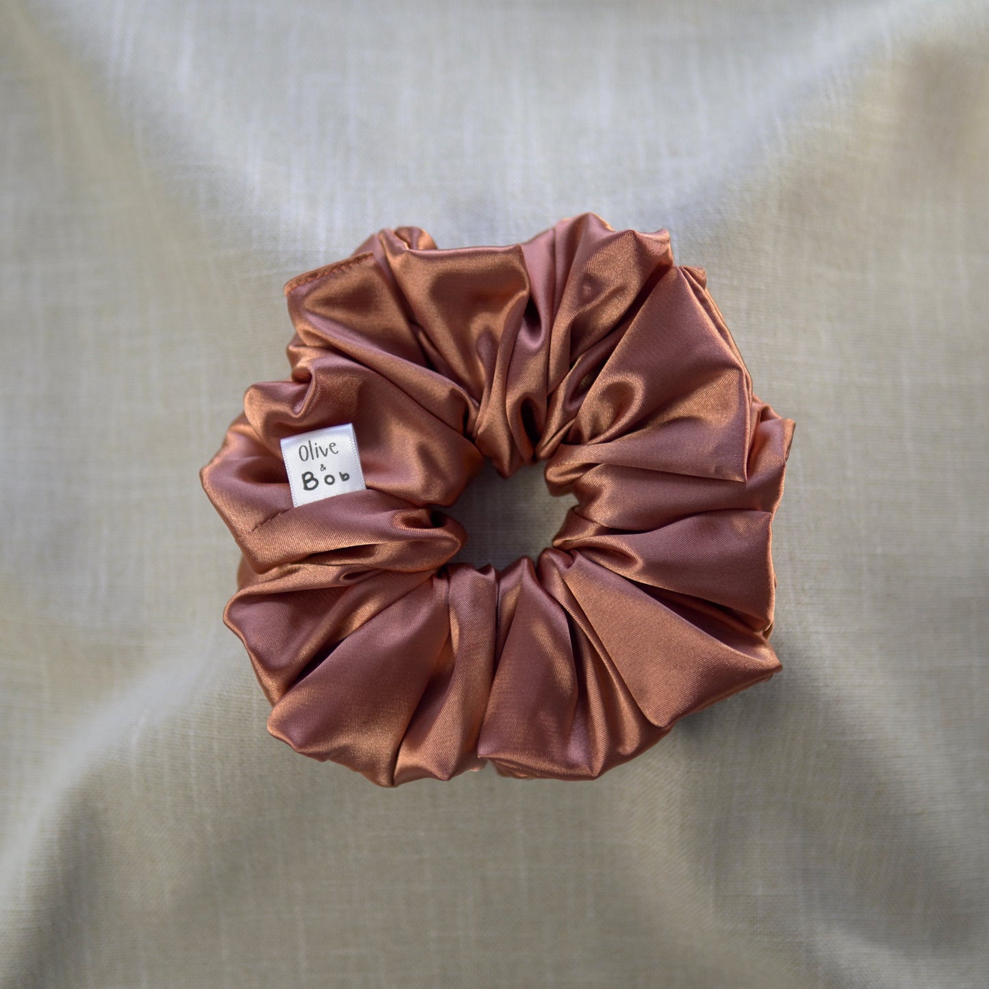 A rusted copper-colored satin scrunchie with a glossy finish, labeled "Olive & Bob," styled on a soft beige fabric background.