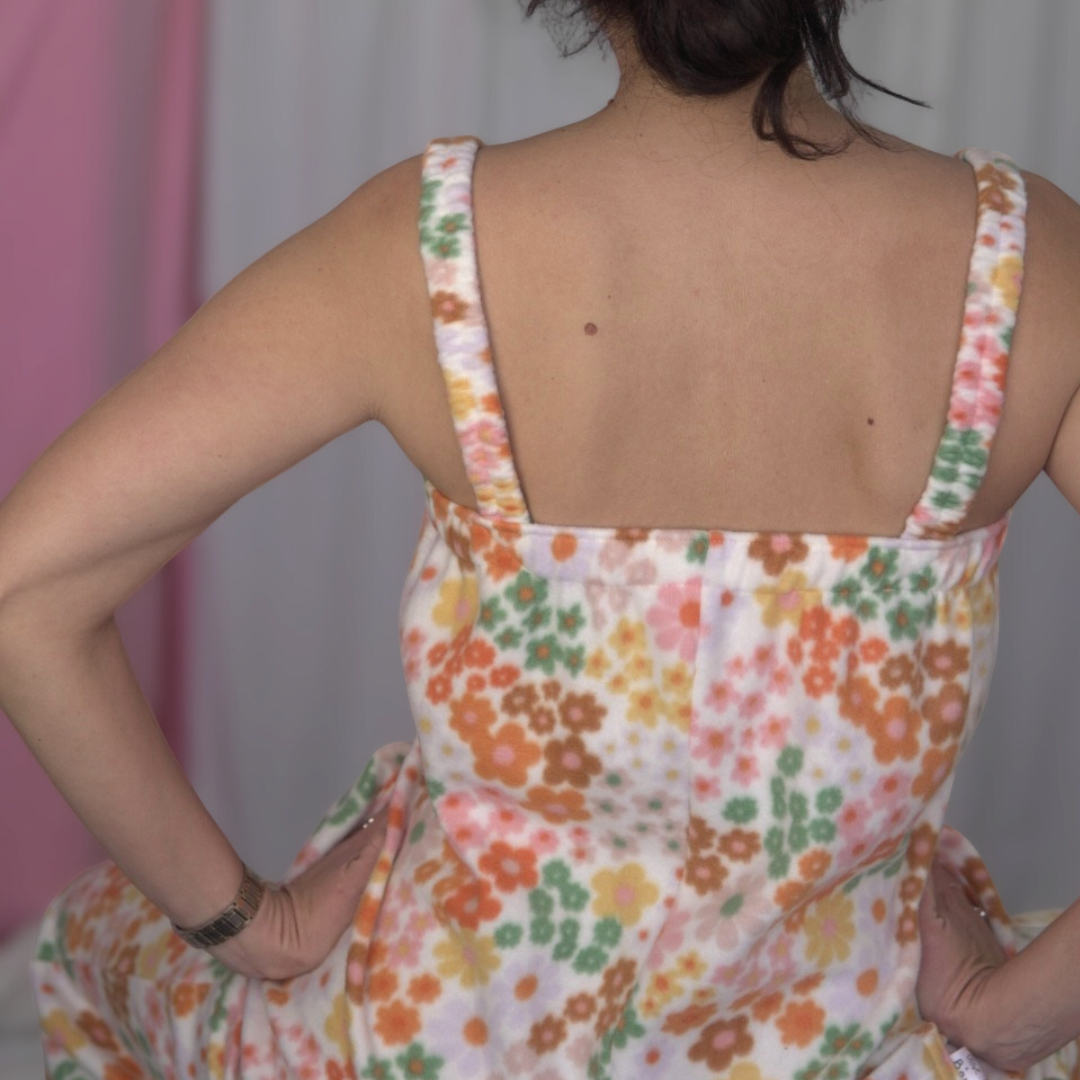 Person wearing a floral fleece jumpsuit with elasticated straps, ankles, and underarm band. The jumpsuit is long and relaxed-fitting, featuring a colorful retro flower print in shades of pink, orange, green, and yellow. Styled with white sneakers, standing in front of a soft pink and white fabric backdrop.