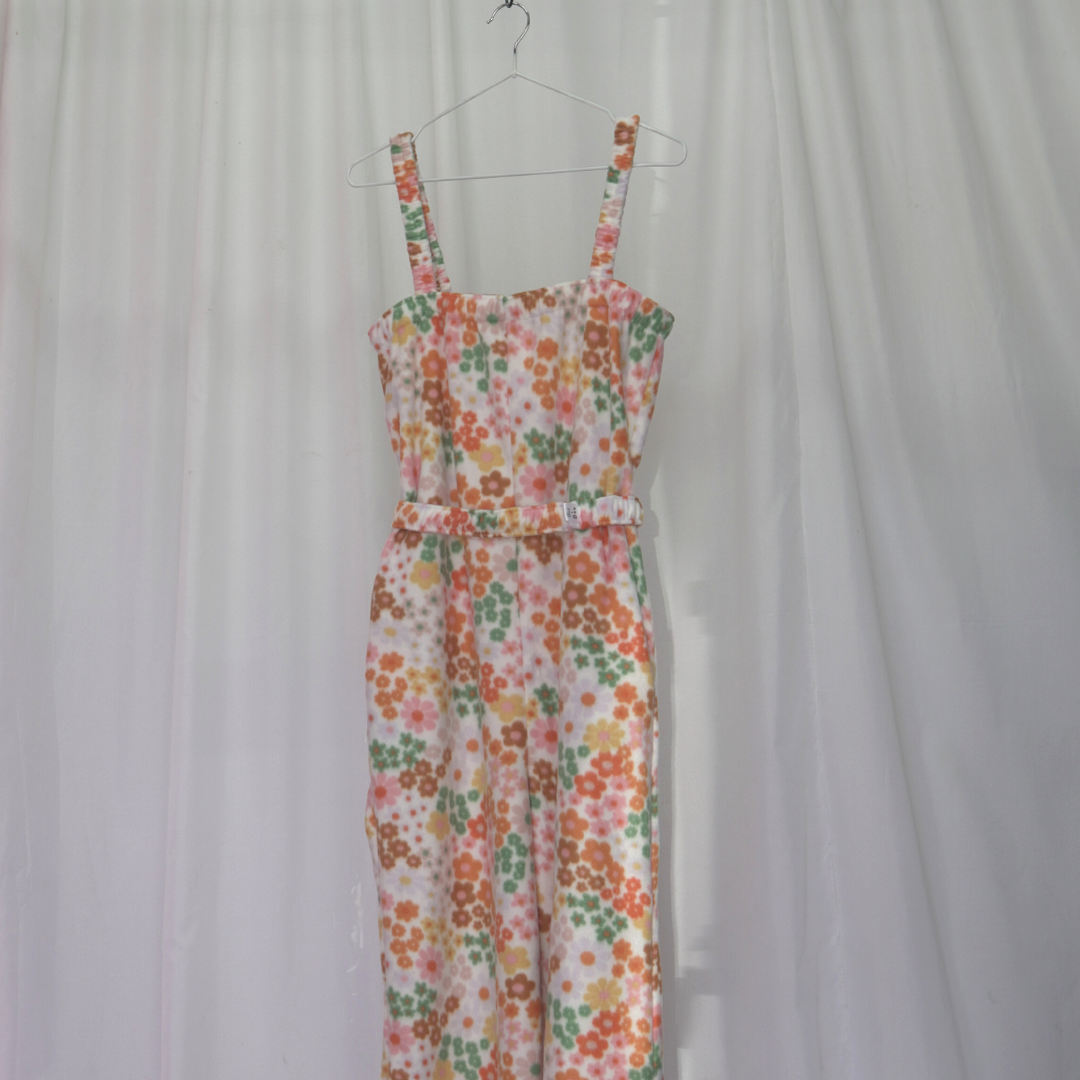 Person wearing a floral fleece jumpsuit with elasticated straps, ankles, and underarm band. The jumpsuit is long and relaxed-fitting, featuring a colorful retro flower print in shades of pink, orange, green, and yellow. Styled with white sneakers, standing in front of a soft pink and white fabric backdrop.
