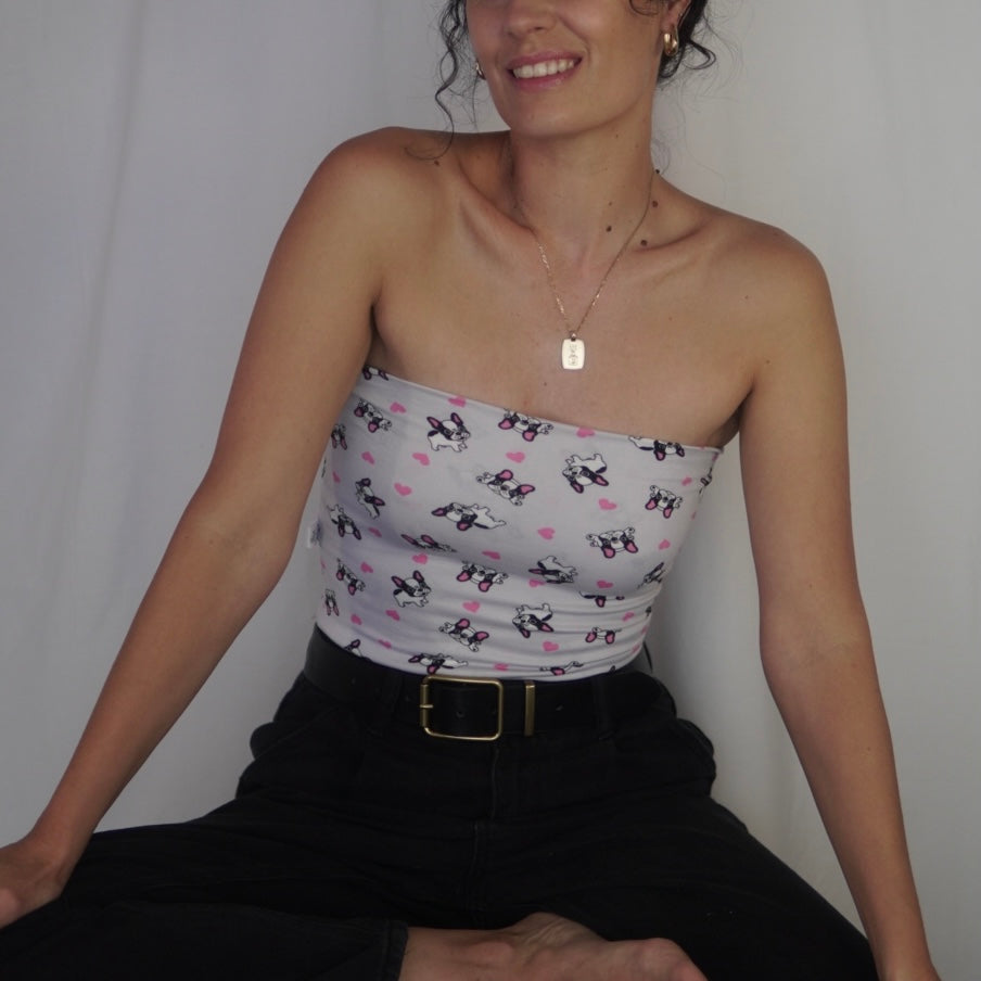 The image shows a woman seated cross-legged, wearing a strapless tube top with a playful French Bulldog and pink heart print on a light gray background. She pairs the top with high-waisted black jeans secured by a black belt with a gold buckle. Her hair is styled in an updo, and she accessorizes with a delicate gold necklace and hoop earrings, smiling softly against a neutral white backdrop.