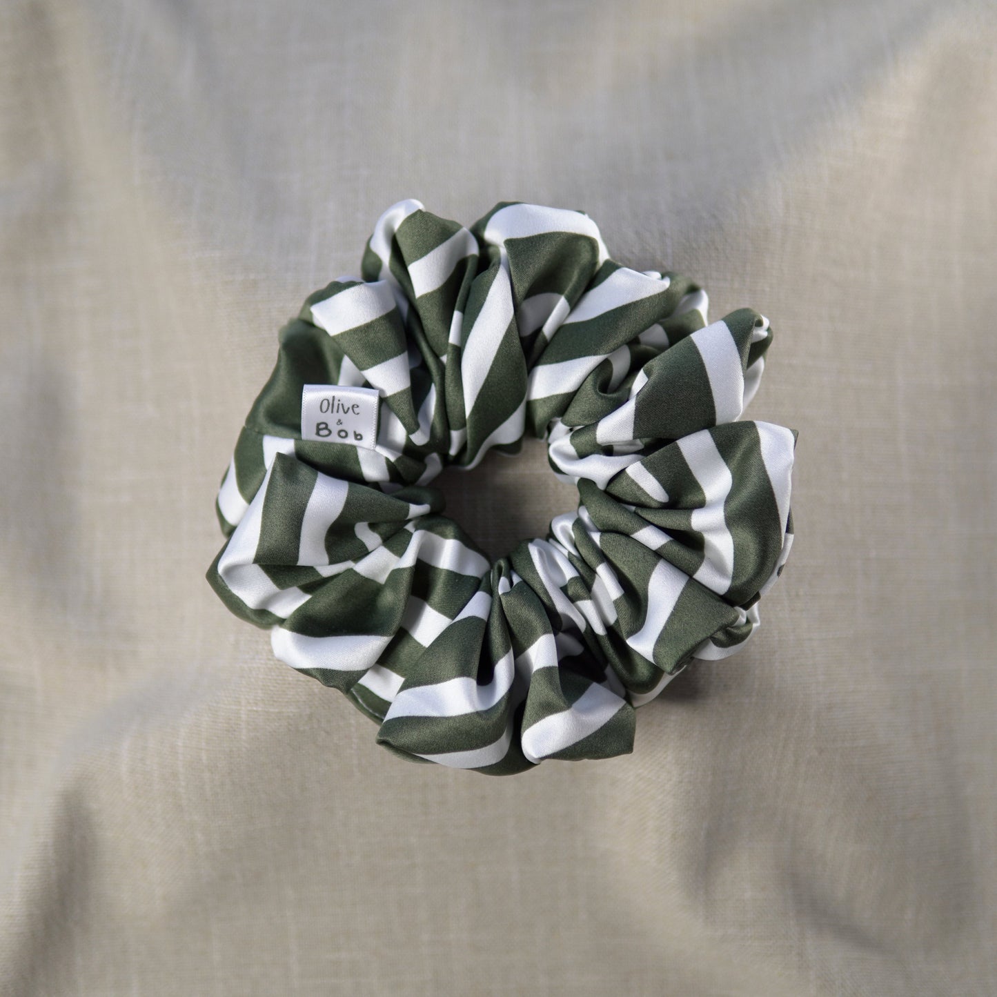 A green and white zebra print scrunchie made from satin-like fabric, displayed on a beige fabric background. The scrunchie features a small 'Olive and Bob' tag.