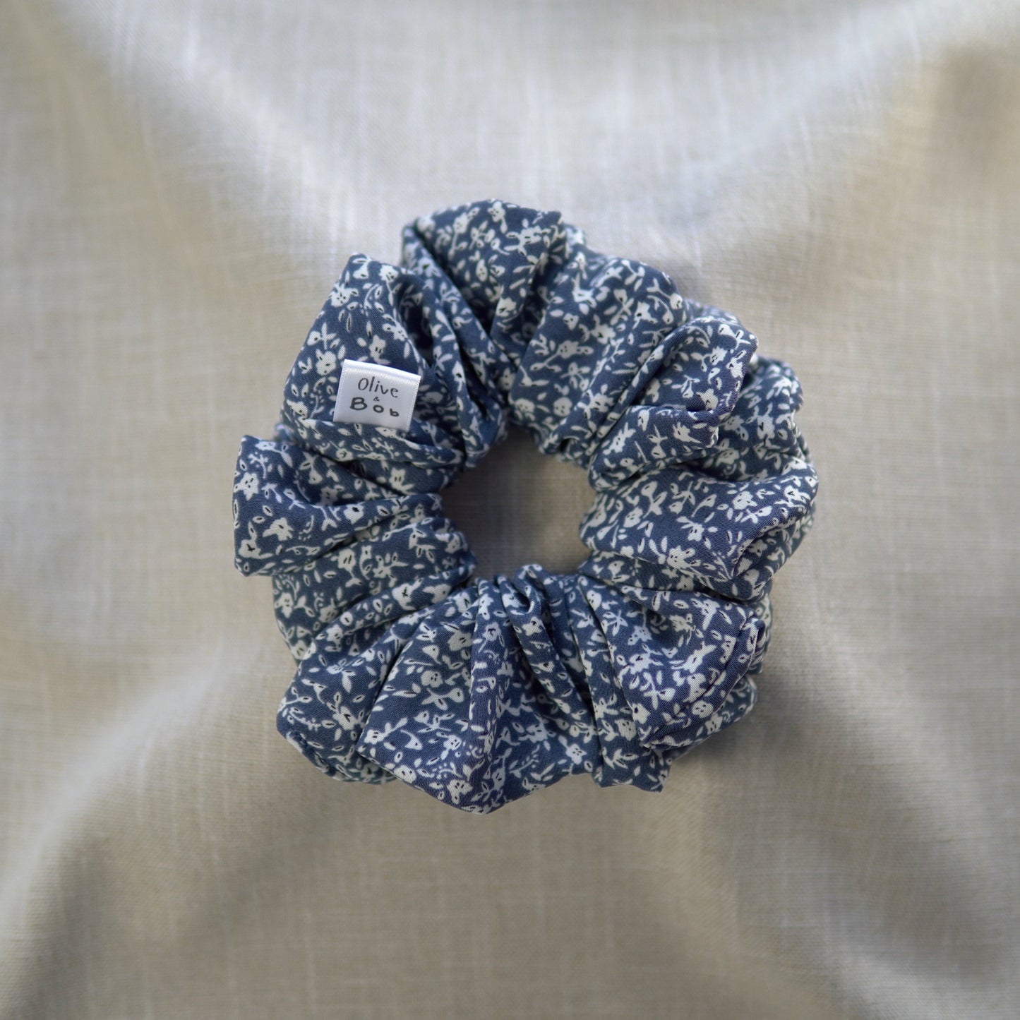 A blue scrunchie made from heavy georgette fabric, featuring a white floral pattern, displayed on a neutral beige fabric background. The scrunchie includes a small 'Olive and Bob' tag.