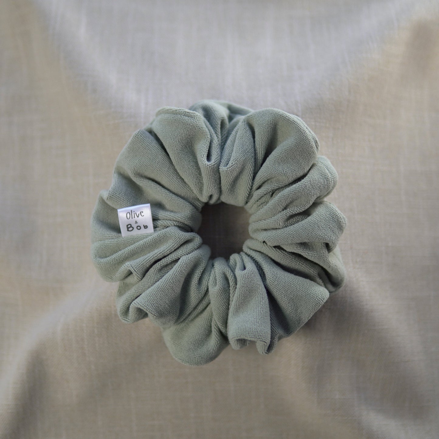 A sage green scrunchie with a soft, towelling-like texture, offering a cozy and stylish hair accessory for everyday use.