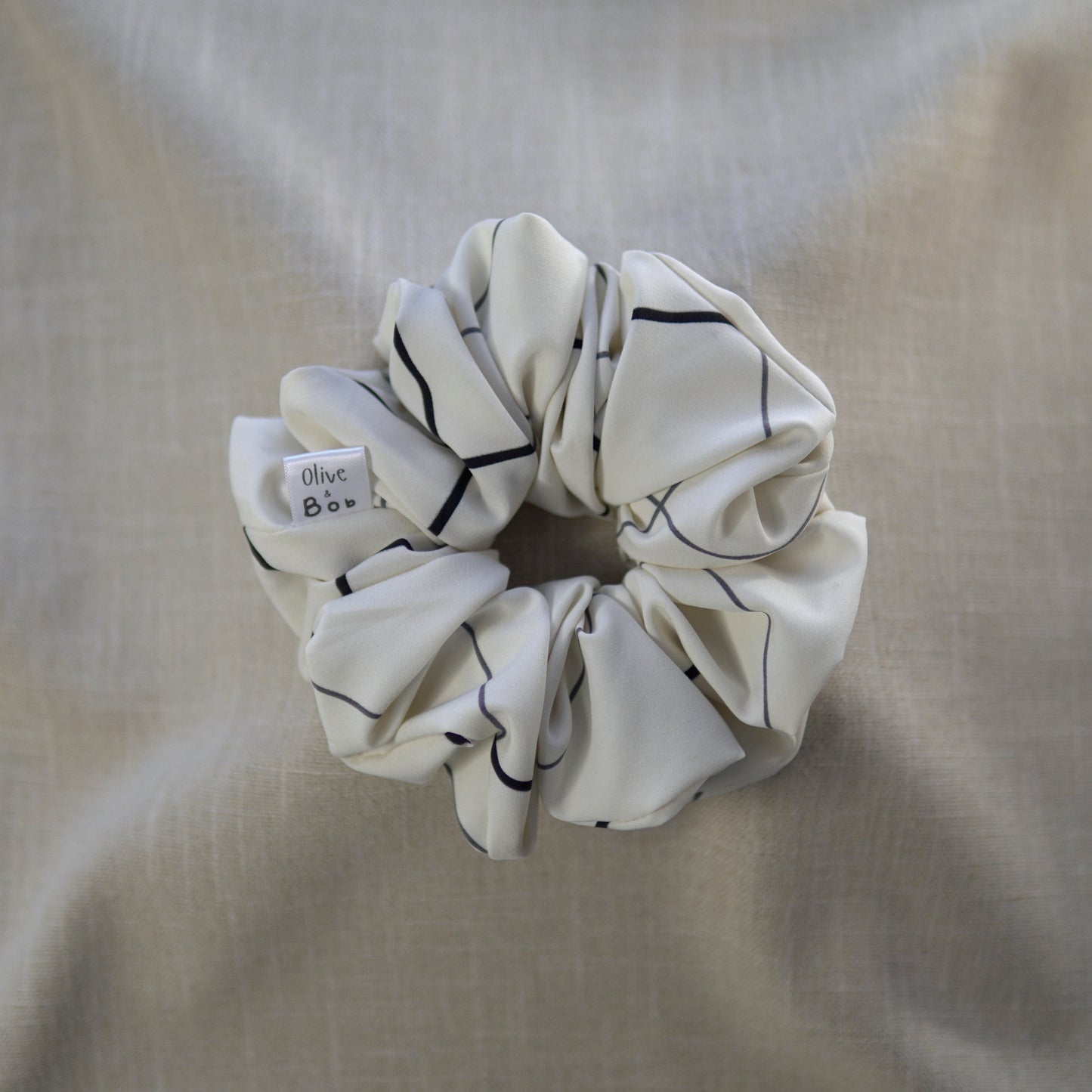A cream-colored scrunchie with soft, chiffon-like fabric featuring a minimalist grid pattern of black horizontal and vertical lines, designed for a lightweight and elegant touch.