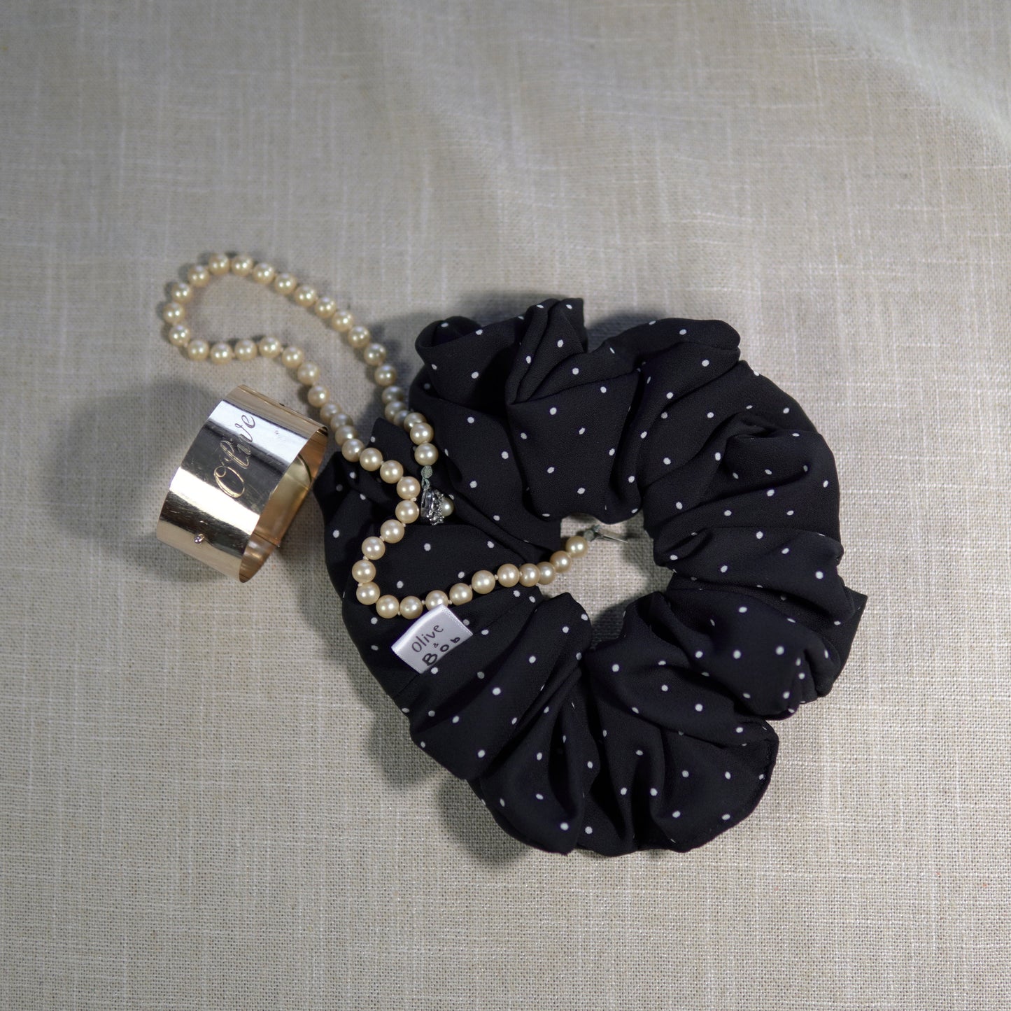 A black chiffon scrunchie adorned with small white polka dots, styled on a neutral fabric backdrop.