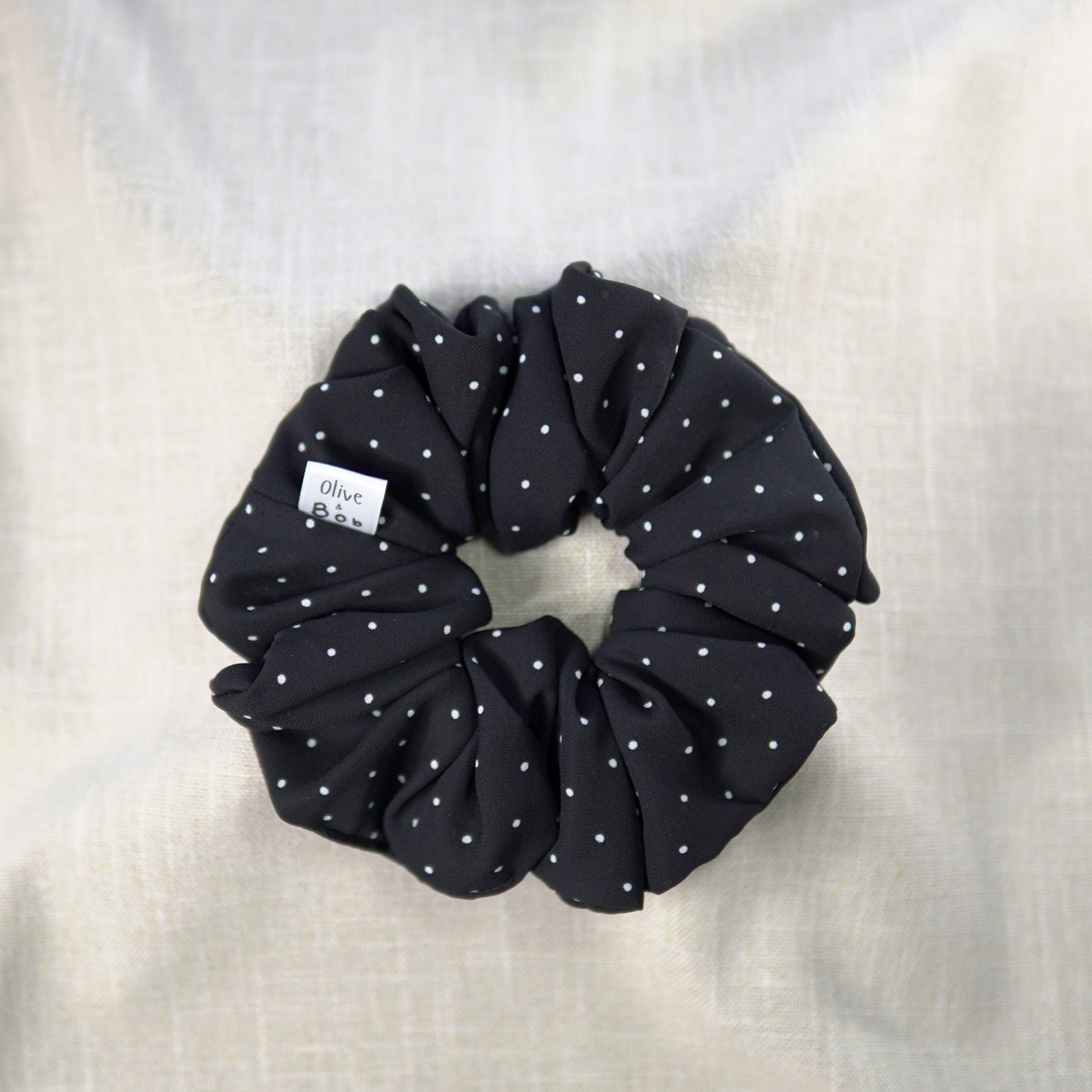 A black chiffon scrunchie adorned with small white polka dots, styled on a neutral fabric backdrop.