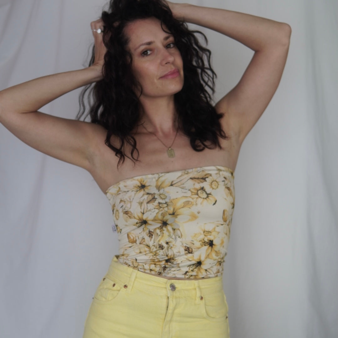 Model wearing a strapless yellow floral tube top with a ruched design, paired with high-waisted yellow pants. The tube top features a vibrant golden floral print, and the model poses with her hands in her hair against a neutral white backdrop