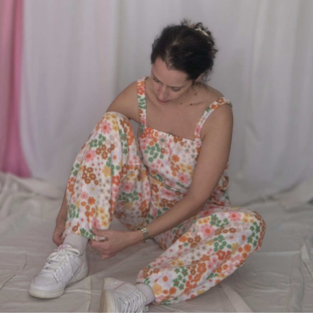 Person wearing a floral fleece jumpsuit with elasticated straps, ankles, and underarm band. The jumpsuit is long and relaxed-fitting, featuring a colorful retro flower print in shades of pink, orange, green, and yellow. Styled with white sneakers, standing in front of a soft pink and white fabric backdrop.
