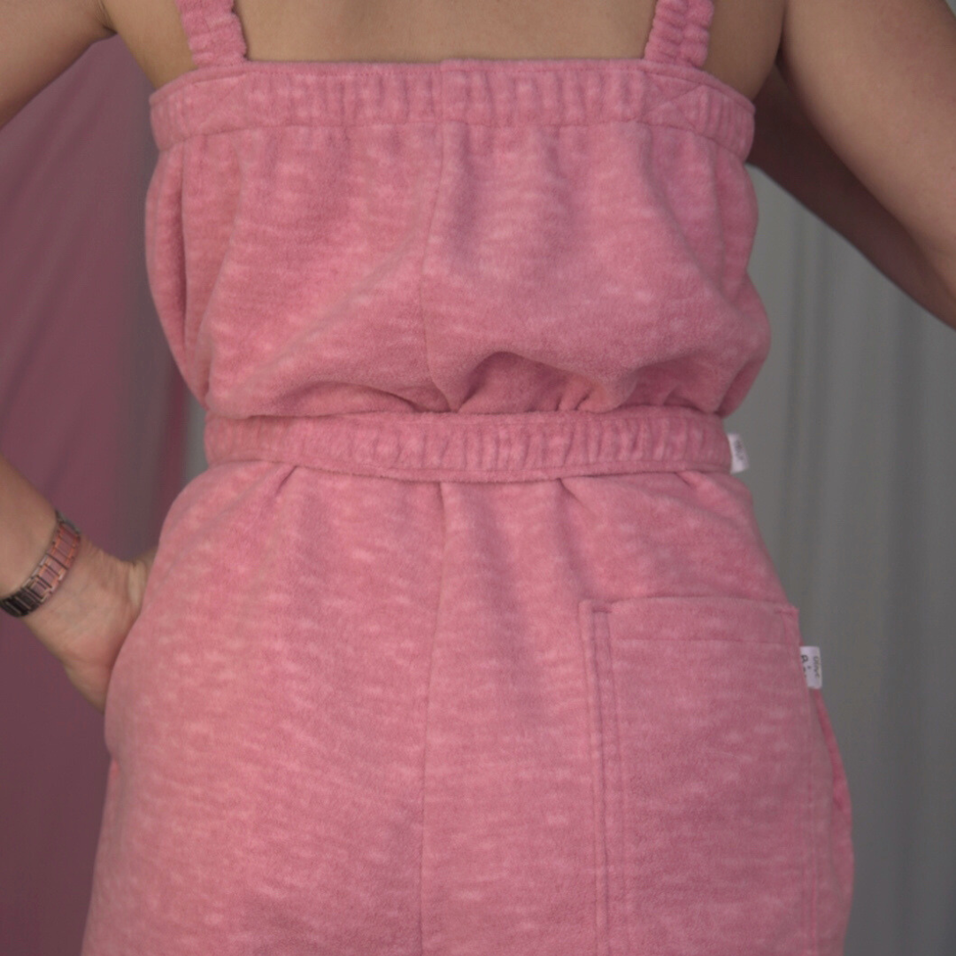 A pink fleece jumpsuit on a model with a soft fabric backdrop. The jumpsuit features elasticated straps, an elastic underarm band, and gathered elastic cuffs at the ankles.