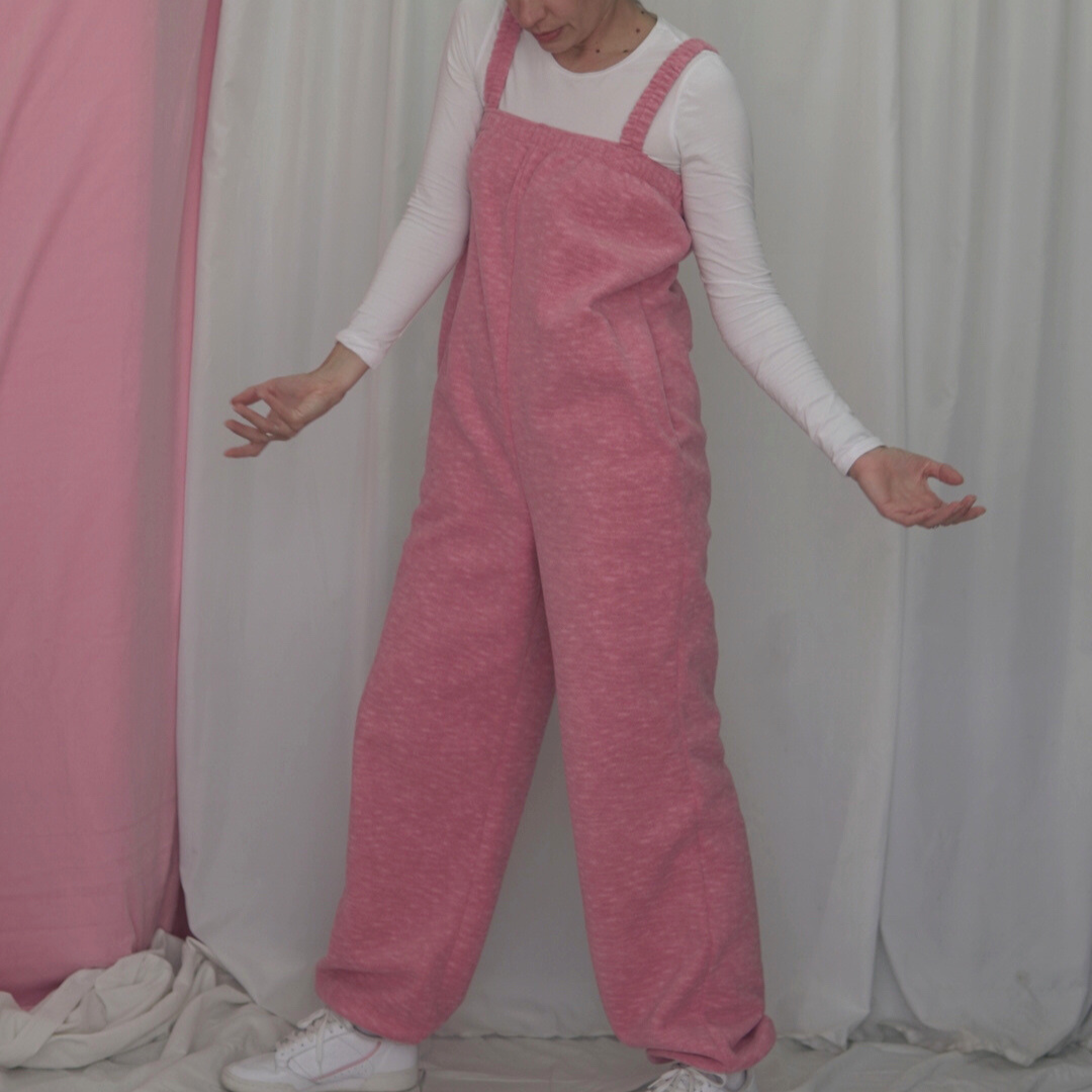 A pink fleece jumpsuit on a model with a soft fabric backdrop. The jumpsuit features elasticated straps, an elastic underarm band, and gathered elastic cuffs at the ankles.