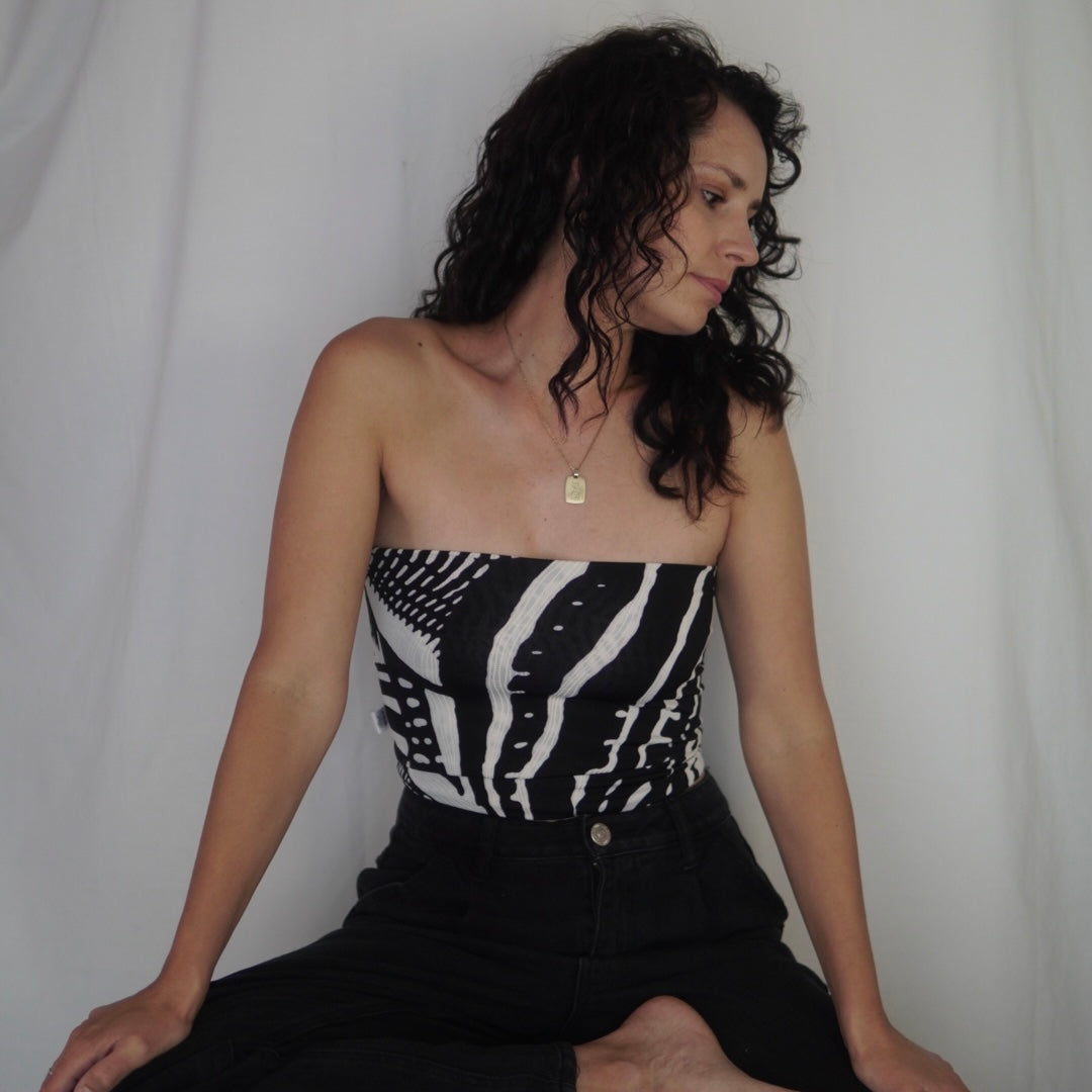 
A woman wearing a strapless tube top with a bold black-and-white abstract pattern, paired with high-waisted black pants. She is seated against a plain white backdrop, gazing thoughtfully to the side, with her curly hair cascading over her shoulders.