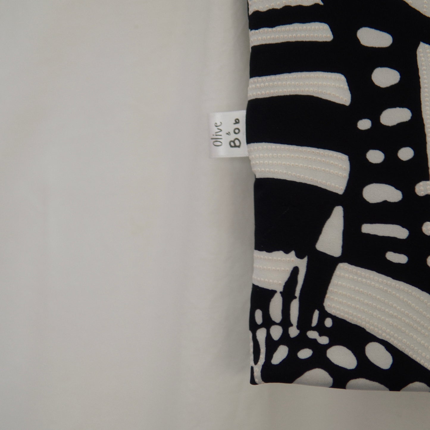 A close-up shot of a black-and-white abstract patterned fabric, emphasizing its intricate dotted and linear design. The "Olive & Bob" label is prominently displayed on the edge, adding a touch of branding against the clean white background.