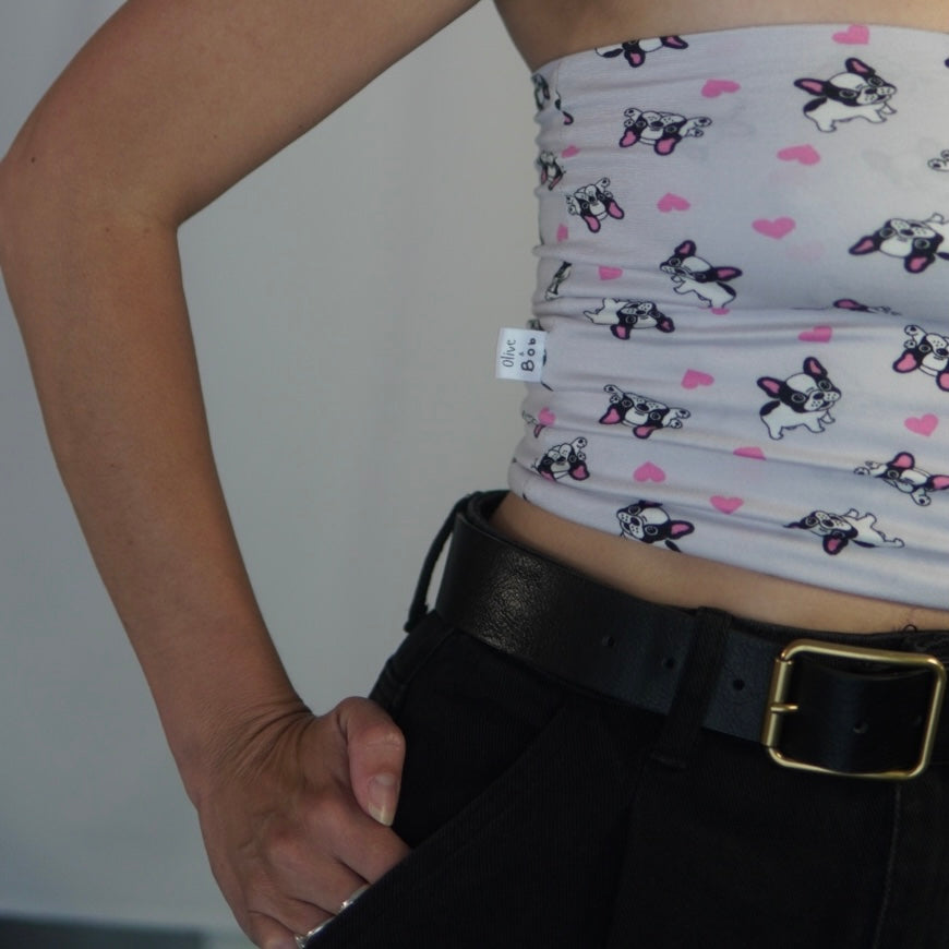 A close-up image of a tube top featuring a playful French Bulldog print with pink heart accents on a light background. The top is paired with black high-waisted pants and a black belt with a gold buckle. A small tag on the side reads "Olive Boo." The person is posing with one hand resting on their hip, emphasizing the details of the outfit.