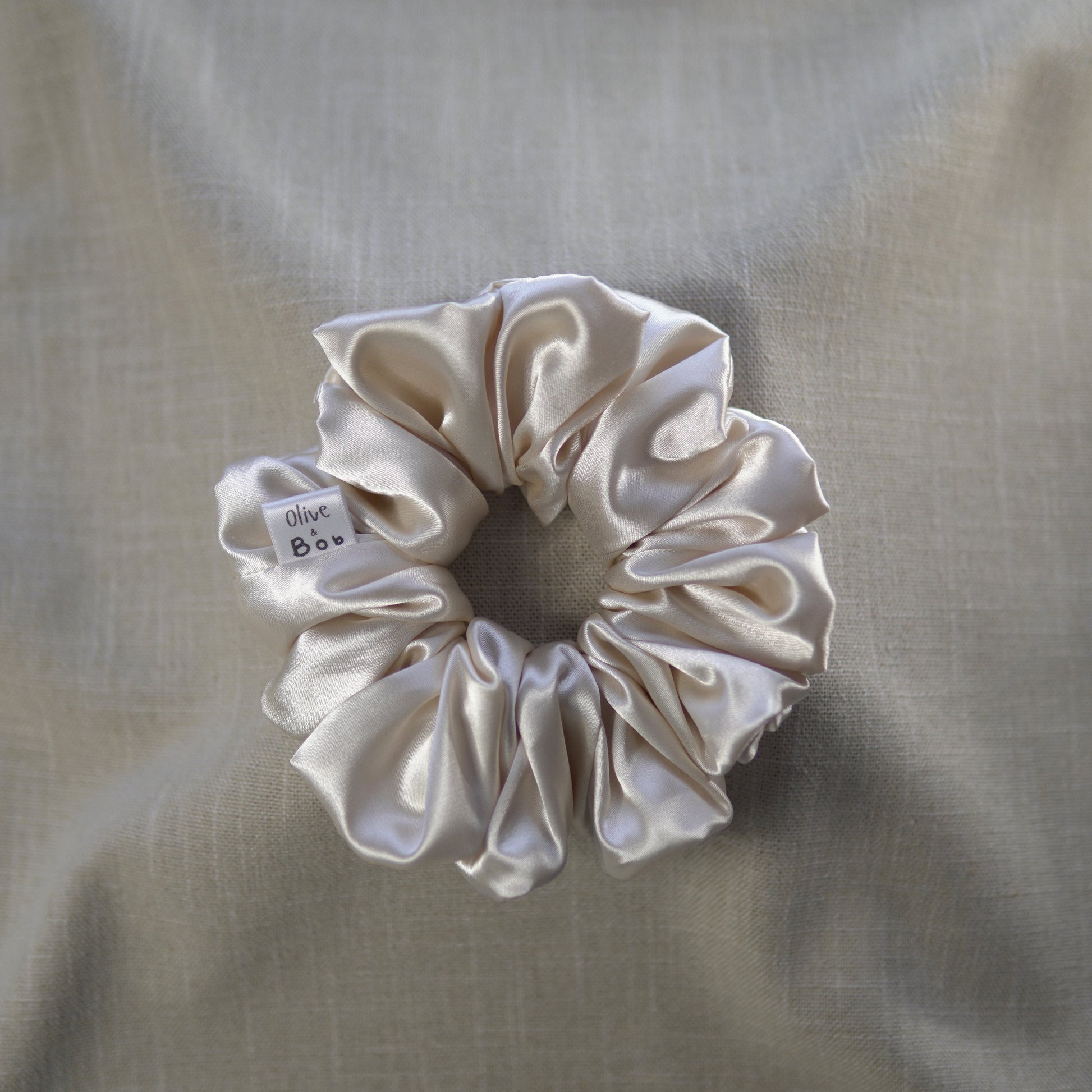 A cream-colored satin scrunchie with a luxurious, smooth texture, designed to add elegance and shine to any hairstyle.