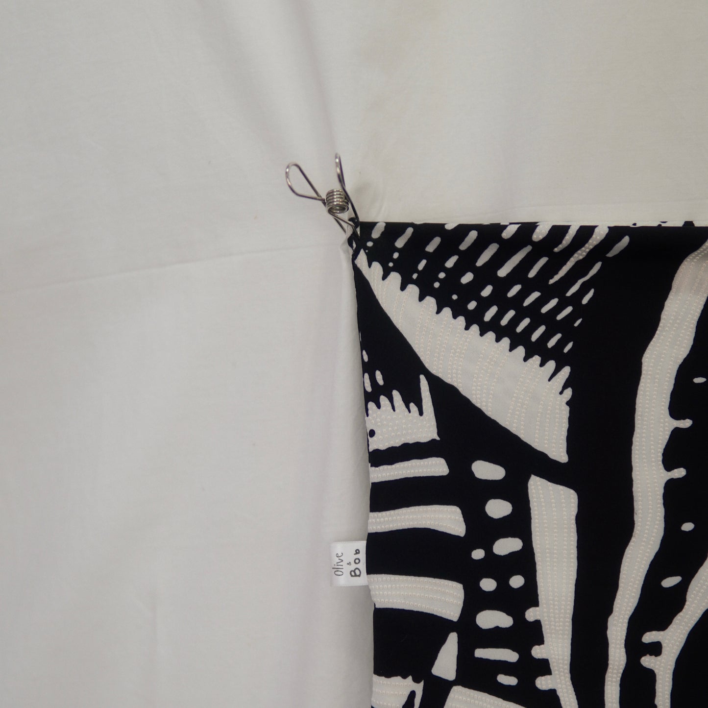 A close-up view of a black-and-white tube top, showcasing its bold abstract pattern with dotted and linear details. The corner of the fabric is pinned, revealing the "Olive & Bob" label, while the minimalist white backdrop enhances the intricate design.