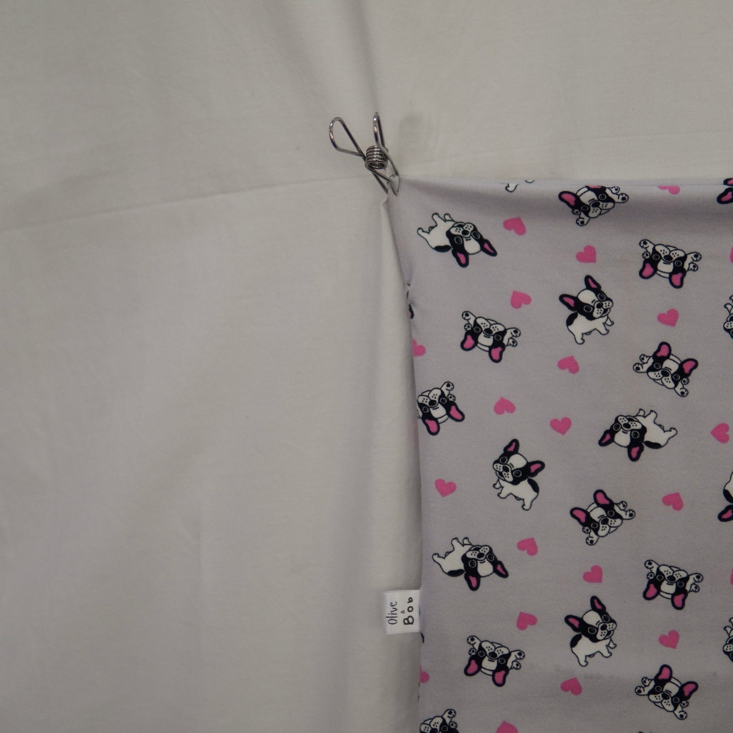 The image shows a close-up of fabric featuring a playful print of French Bulldogs and pink hearts on a light gray background. A small "Olive & Bob" tag is visible on the edge of the fabric, which is clipped at the top against a plain white backdrop.