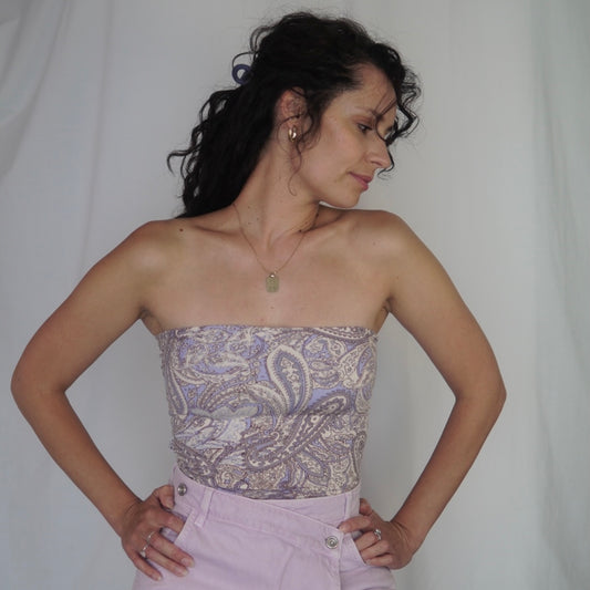 his image showcases a woman modeling a strapless tube top featuring an elegant paisley design in soft lavender, cream, and gray tones. The top is paired with high-waisted lavender pants that complement the outfit's color palette. Her pose, with hands on her hips, highlights the garment's fit and style, while her minimal jewelry, including a pendant necklace and hoop earrings, adds a refined touch. The background is a neutral drape, emphasizing the details of the attire.