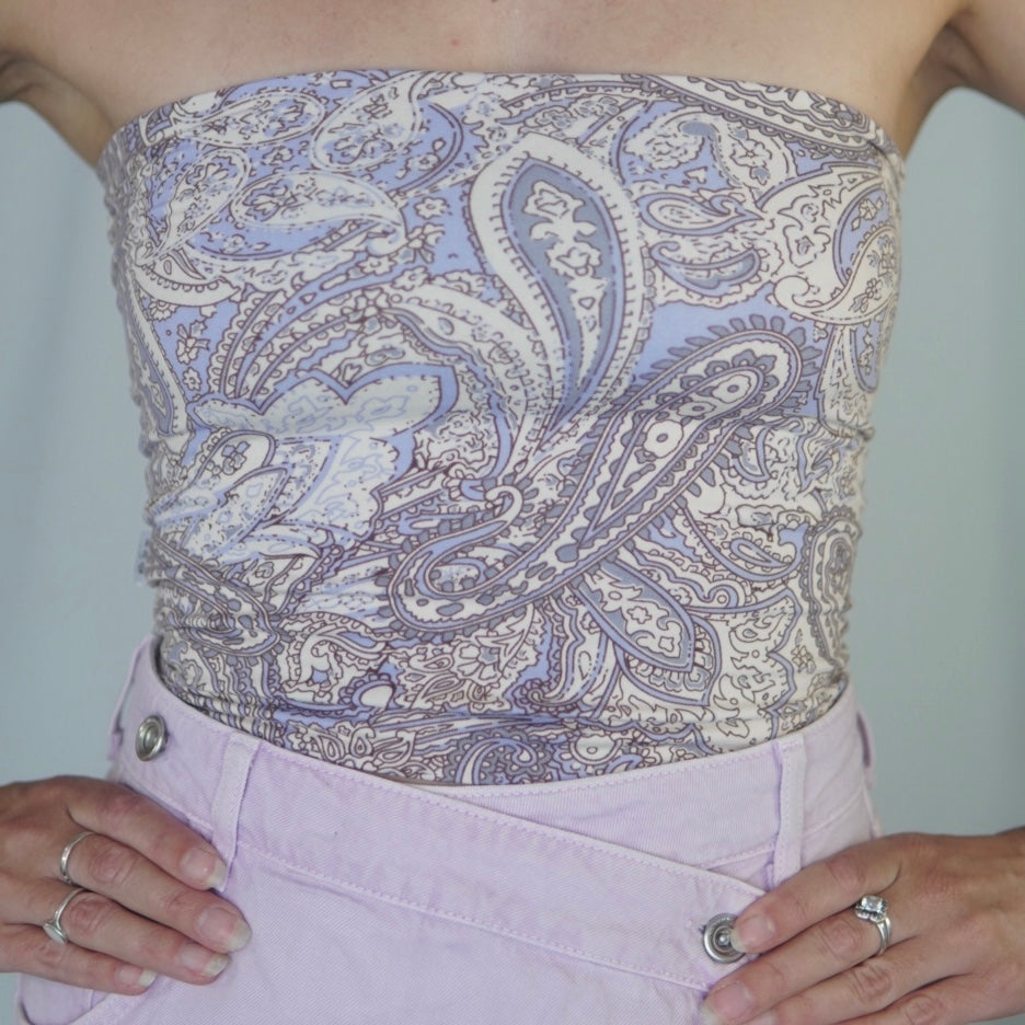 A close-up of a strapless tube top with an intricate paisley pattern in shades of lavender, cream, and gray. The top is paired with high-waisted pastel lavender pants, with the wearer’s hands resting on the waistband. The overall look has a boho-inspired aesthetic.