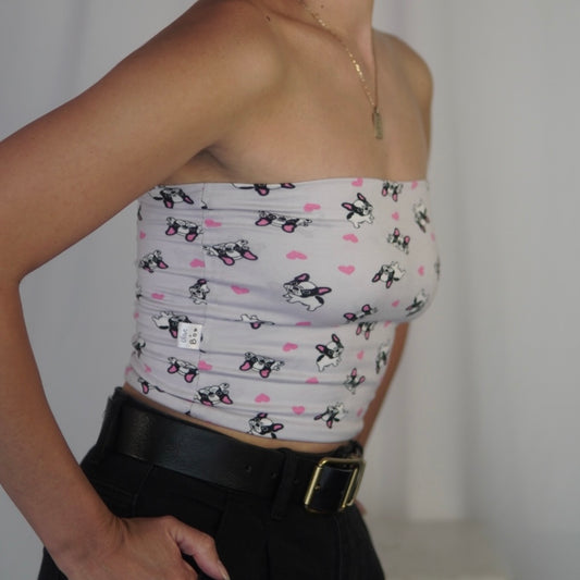 A close-up image of a tube top featuring a playful French Bulldog print with pink heart accents on a light background. The top is paired with black high-waisted pants and a black belt with a gold buckle. A small tag on the side reads "Olive & Bob." The person is posing with one hand resting on their hip, emphasizing the details of the outfit.