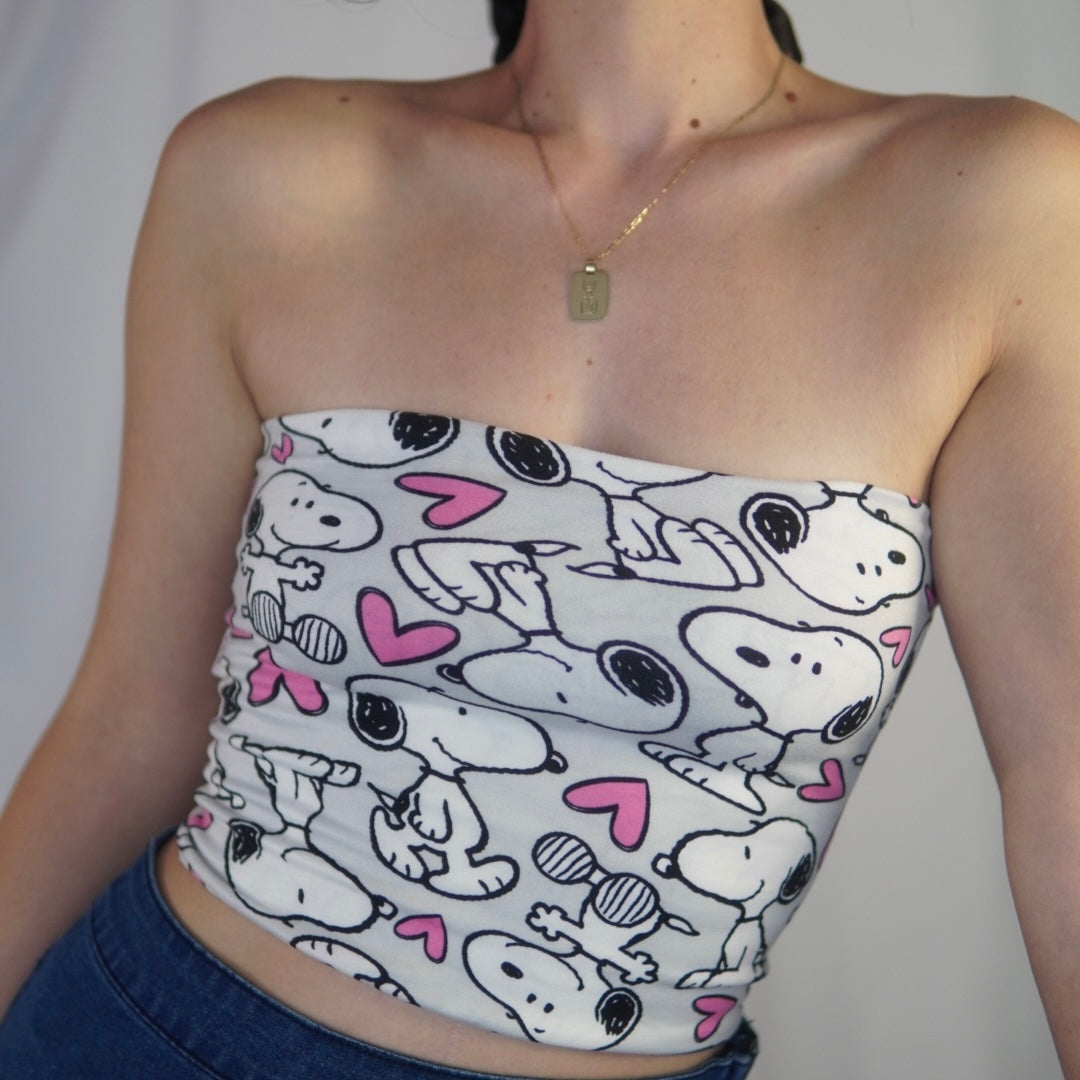 Close-up of a woman wearing a strapless top with a playful Snoopy and pink hearts pattern. She has a gold necklace with a rectangular pendant and is paired with high-waisted blue jeans. The background is plain and softly lit