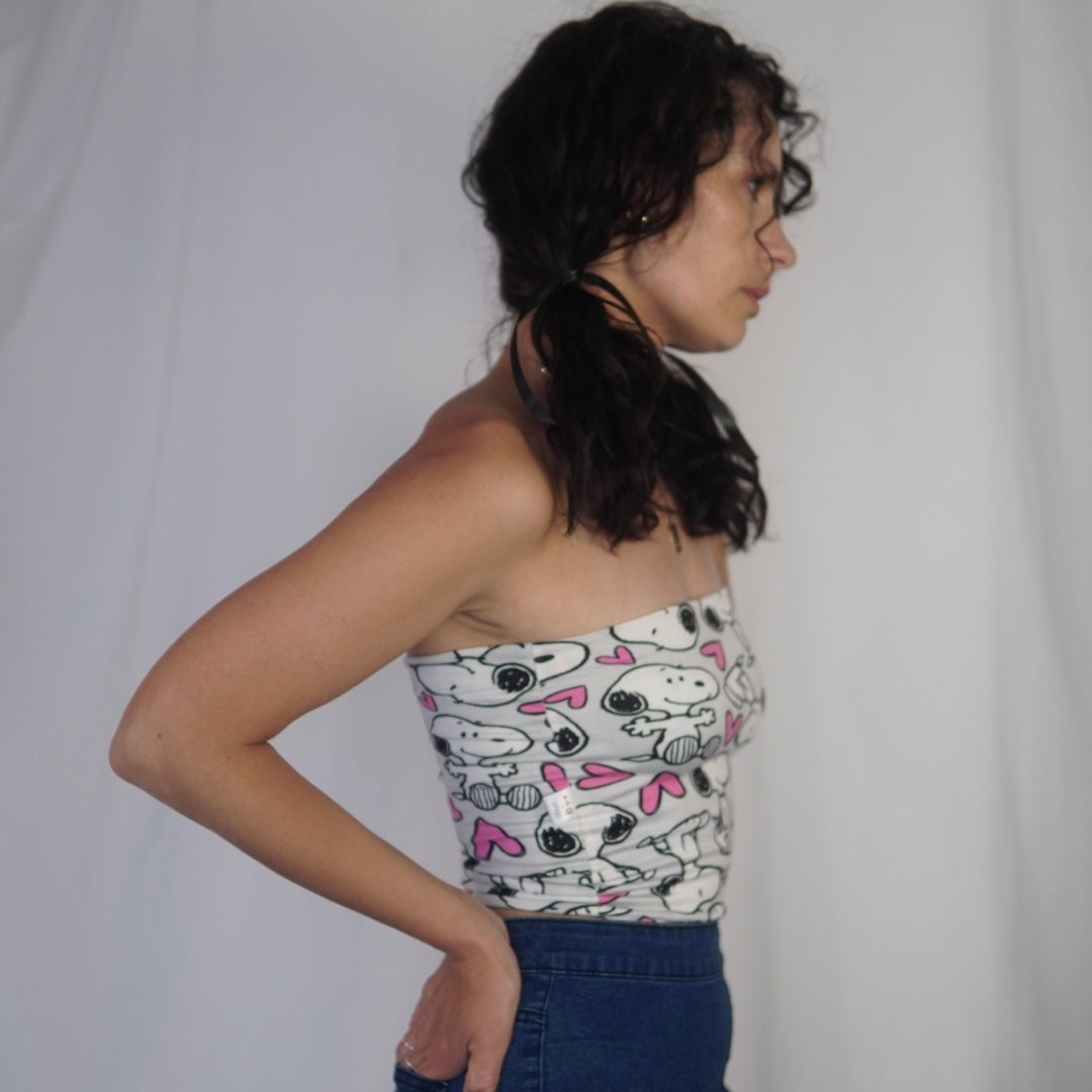 A woman in profile view standing against a plain white background. She has dark curly hair styled in pigtails and is wearing a strapless top featuring a playful design of Snoopy characters and pink hearts. She pairs it with high-waisted blue jeans and has her hands resting on her hips