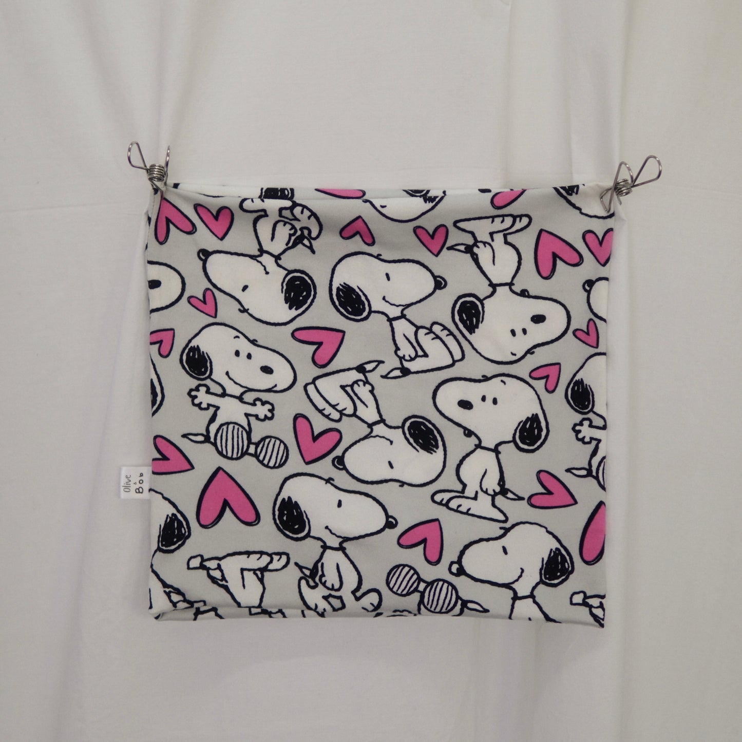 A rectangular piece of fabric displayed against a white background. The fabric features a repeating pattern of Snoopy illustrations in various poses, surrounded by pink hearts on a light gray background. The corners of the fabric are pinned up with metal clips