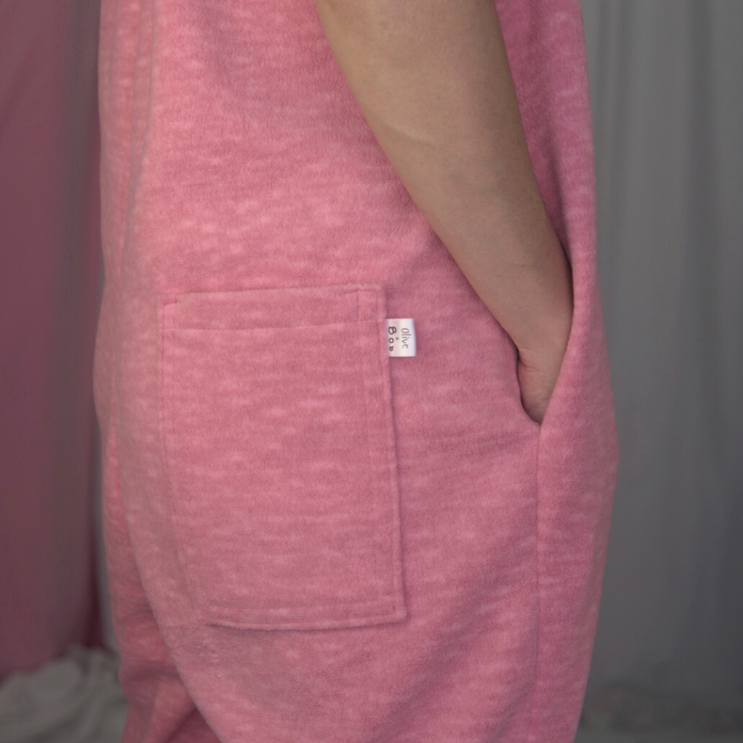 A pink fleece jumpsuit on a model with a soft fabric backdrop. The jumpsuit features elasticated straps, an elastic underarm band, and gathered elastic cuffs at the ankles.