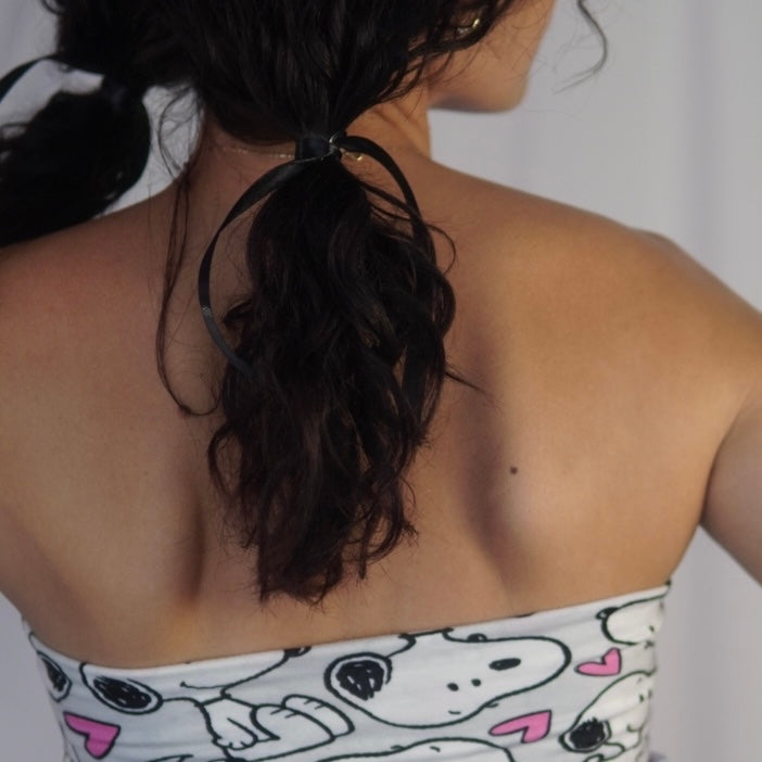 Close-up view of a woman's back, showing her dark curly hair tied into pigtails with black ribbons. She is wearing a strapless top featuring a playful Snoopy pattern with pink hearts on a light background