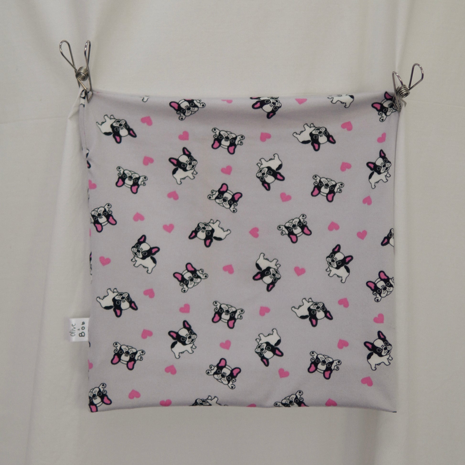 The image displays a close-up of a tube top stretched and clipped at the corners. The top features a playful pattern of French Bulldogs in black and white with pink accents, interspersed with pink hearts, all set against a light gray background. A small white tag with "Olive & Bob" branding is visible on the left edge. The top is displayed against a neutral white fabric backdrop.